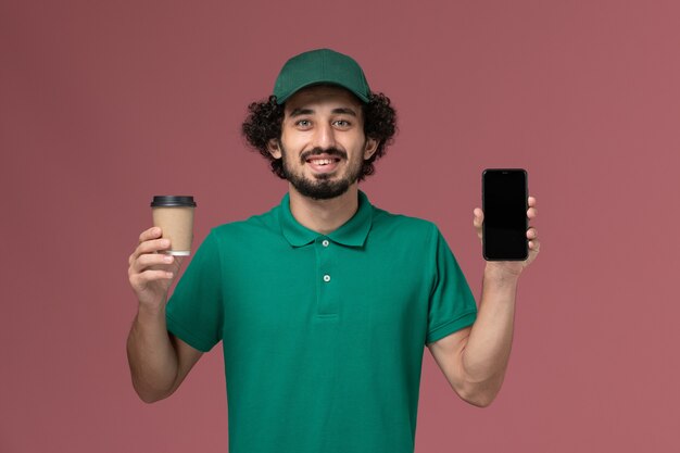 Corriere maschio di vista frontale in uniforme verde e tazza di caffè di consegna della tenuta del capo con il telefono sul lavoro dell'azienda di servizio di consegna uniforme del fondo rosa