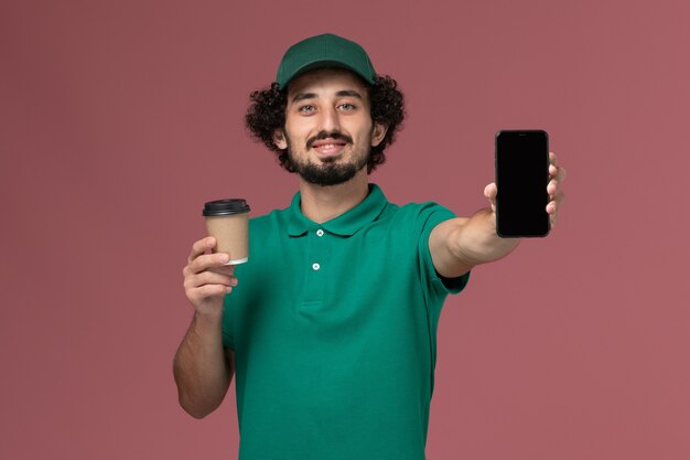 Corriere maschio di vista frontale in uniforme verde e tazza di caffè di consegna della tenuta del capo con il telefono sul servizio di consegna uniforme del fondo rosa