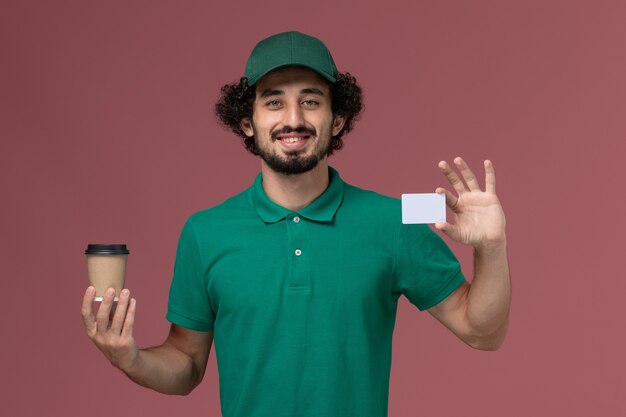 Corriere maschio di vista frontale in uniforme verde e tazza di caffè di consegna della tenuta del capo con la carta sul lavoratore di lavoro dell'azienda di servizio di consegna uniforme del fondo rosa