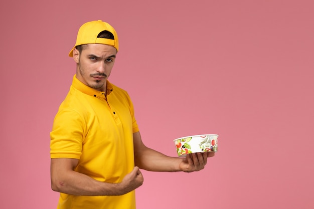 Corriere maschio di vista frontale nella ciotola di consegna della tenuta della tenuta uniforme gialla e posa su fondo rosa.