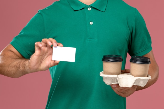 Corriere maschio di vista ravvicinata anteriore in uniforme verde che tiene tazze di caffè marrone di consegna e carta di plastica bianca sul fondo rosa dello scrittorio