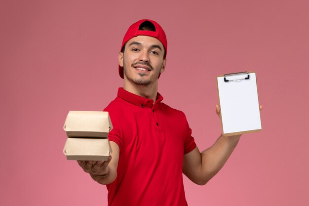 Corriere maschio giovane vista frontale in mantello rosso uniforme che tiene blocco note e piccolo pacchetto con cibo su sfondo rosa.