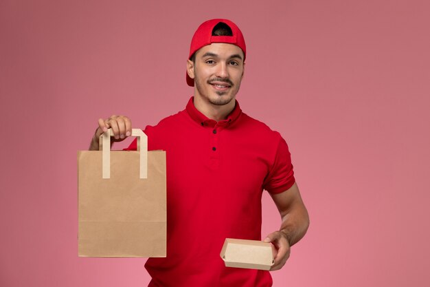 Corriere maschio giovane vista frontale in mantello rosso uniforme che tiene due diversi pacchetti di cibo con sorriso su sfondo rosa.