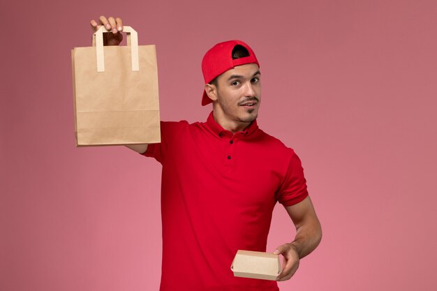 Corriere maschio giovane vista frontale in mantello rosso uniforme che tiene due diversi pacchetti di cibo sullo sfondo rosa.