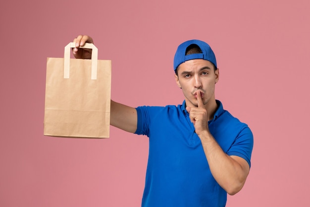 Corriere maschio giovane vista frontale in uniforme blu e mantello con pacco di carta di consegna sulle mani sulla parete rosa