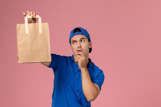 Corriere maschio giovane vista frontale in uniforme blu e mantello con pacco di carta di consegna sulle sue mani pensando sulla parete rosa