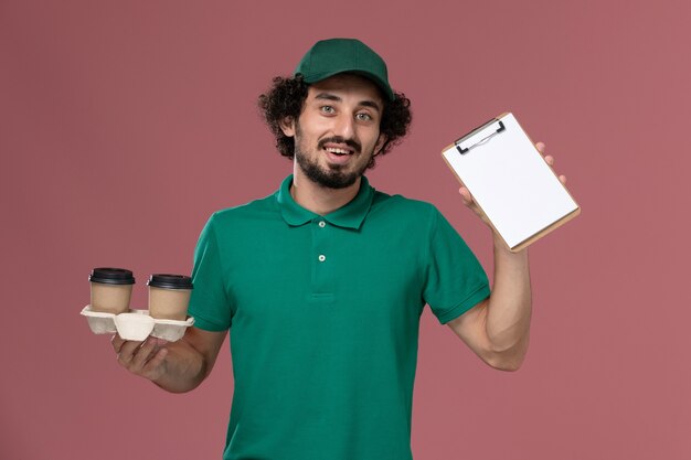 Corriere maschio giovane vista frontale in uniforme verde e mantello che tiene le tazze di caffè di consegna e blocco note sulla consegna uniforme di lavoro di servizio lavoratore maschio sfondo rosa