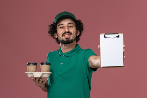 Corriere maschio giovane vista frontale in uniforme verde e mantello che tiene le tazze di caffè di consegna e blocco note sulla consegna uniforme di lavoro di servizio sfondo rosa chiaro