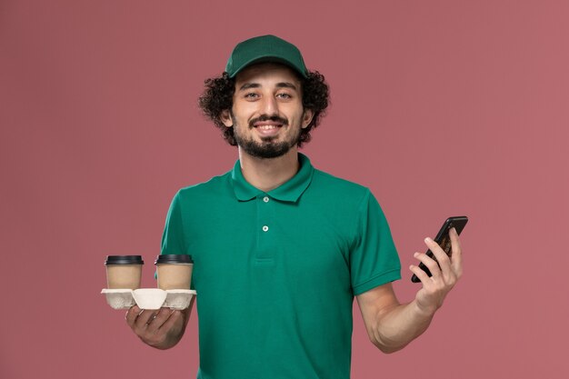 Corriere maschio giovane vista frontale in uniforme verde e mantello che tiene le tazze di caffè di consegna e il suo telefono che sorride sull'operaio di consegna uniforme di lavoro di sfondo rosa