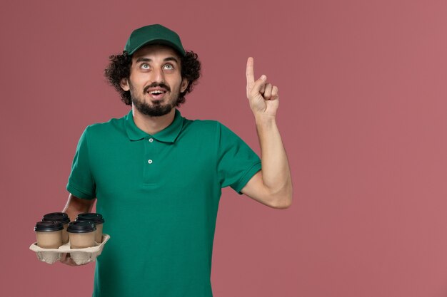 Corriere maschio giovane vista frontale in uniforme verde e mantello che tiene tazze di caffè consegna marrone in posa e pensando sul lavoro di operaio consegna uniforme servizio sfondo rosa