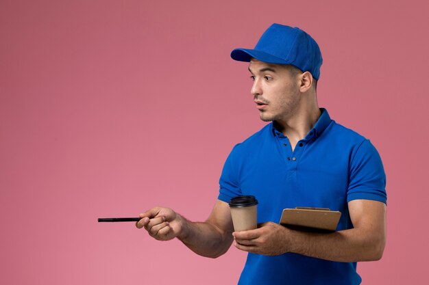 corriere maschio in uniforme blu che tiene la penna del blocco note della tazza di caffè sul rosa, consegna del servizio uniforme del lavoratore