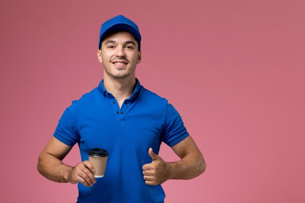 corriere maschio in uniforme blu che tiene tazza di caffè e sorridente sul rosa, consegna del servizio uniforme operaio