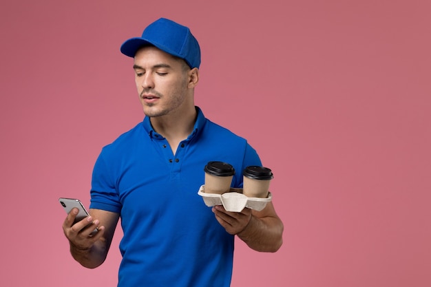 corriere maschio in uniforme blu che tiene tazze di caffè e usando il suo telefono sulla consegna del lavoro di servizio rosa, uniforme