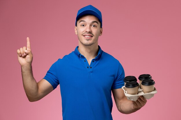 corriere maschio in uniforme blu che tiene tazze di caffè marroni con il dito alzato sul rosa, consegna del servizio uniforme operaio