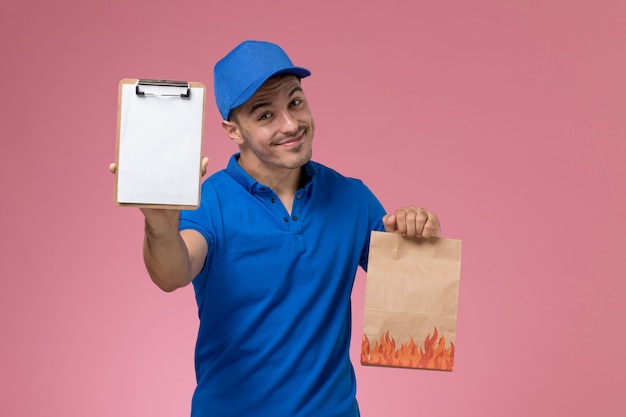 Corriere maschio vista frontale in uniforme blu che tiene il pacchetto di carta alimentare e blocco note sulla parete rosa, consegna del lavoro di servizio uniforme