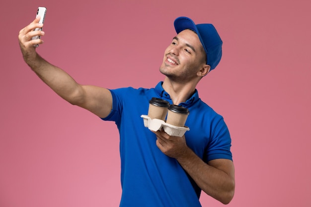 Corriere maschio vista frontale in uniforme blu prendendo selfie con tazze di caffè sul muro rosa, consegna del lavoro di servizio uniforme