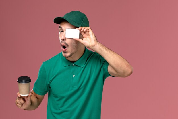 Corriere maschio vista frontale in uniforme verde che tiene la tazza di caffè di consegna con carta bianca su sfondo rosa chiaro