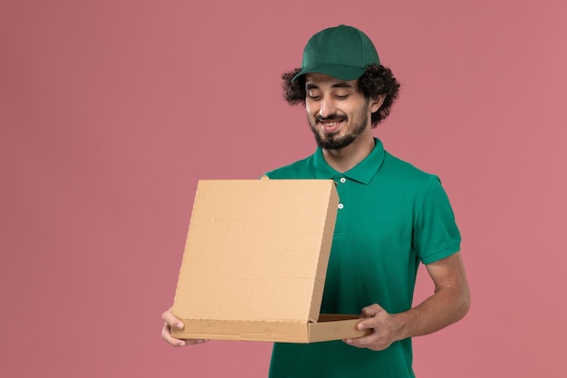 Corriere maschio vista frontale in uniforme verde e mantello che tiene scatola di cibo aprendolo su sfondo rosa servizio di consegna uniforme operaio maschio