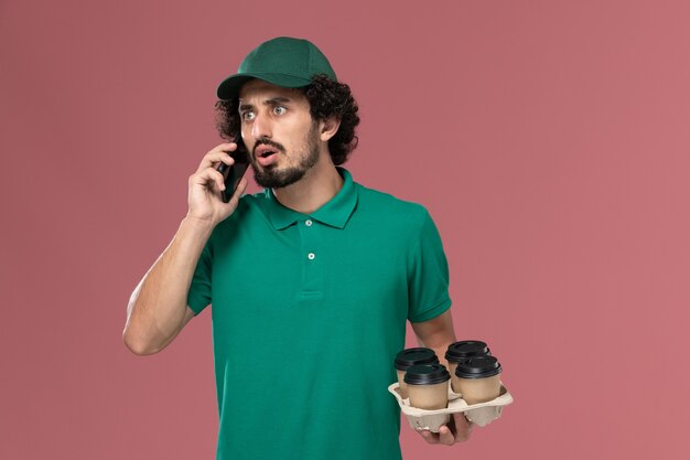 Corriere maschio vista frontale in uniforme verde e mantello che tiene tazze di caffè e parlando al telefono su sfondo rosa chiaro servizio uniforme consegna lavoro maschile