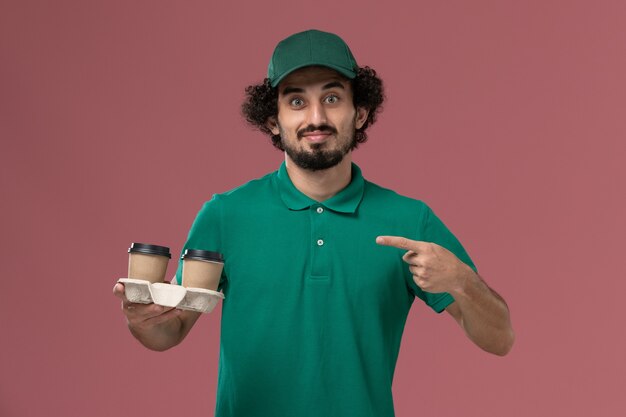 Corriere maschio vista frontale in uniforme verde e mantello che tiene tazze di caffè sullo sfondo rosa servizio di consegna uniforme lavoro lavoratore lavoro maschile