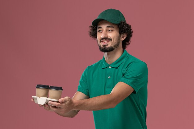 Corriere maschio vista frontale in uniforme verde e mantello che tiene tazze di caffè sullo sfondo rosa uniforme servizio di consegna lavoro azienda maschile