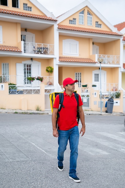 Corriere sicuro che consegna l'ordine e lavora nel servizio postale. Fattorino che indossa jeans, berretto rosso e camicia, portando uno zaino termico giallo. Servizio di consegna di cibo e concetto di acquisto online