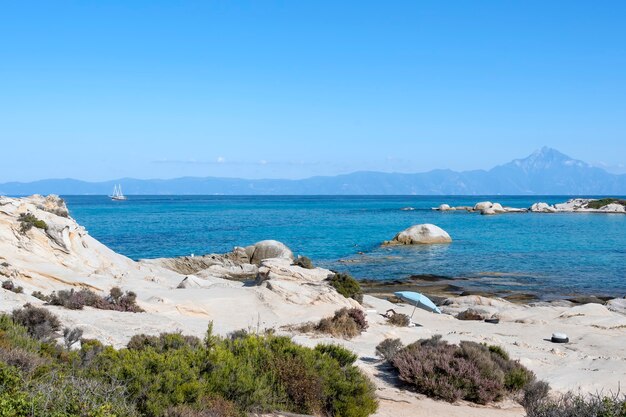 Costa del Mar Egeo con persone che nuotano, rocce sull'acqua e terra con la barca in lontananza, vegetazione in primo piano, acqua blu, Grecia