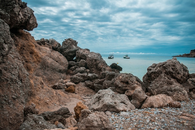 Costa piena di rocce, alcune navi in mare e il cielo nuvoloso