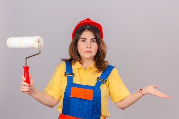 Costruttore donna in uniforme da costruzione e casco di sicurezza in piedi con il rullo di vernice in mano scrollando le spalle allargando le mani non capendo cosa è successo espressione confusa e confusa sopra