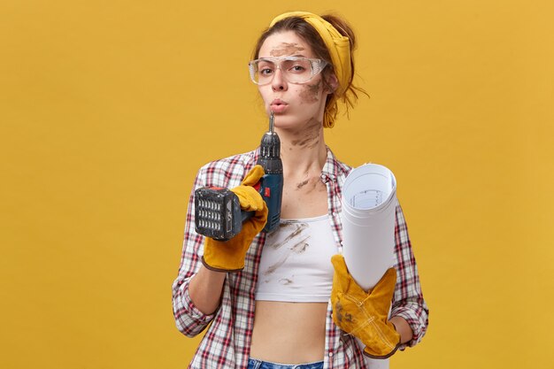 Costruttore femminile sicuro di sé che indossa occhiali protettivi, top bianco e camicia a scacchi, guanti protettivi che tengono trapano e documenti sporchi dopo un duro lavoro isolato sopra il muro giallo. Manutenzione