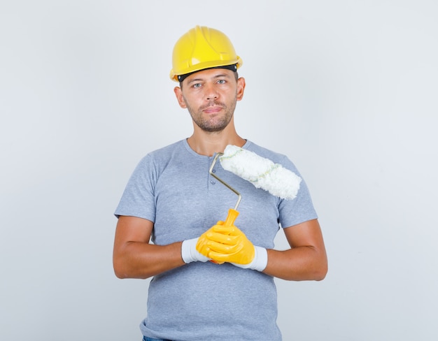 Costruttore maschio in t-shirt, casco, guanti che tengono rullo di vernice, vista frontale.