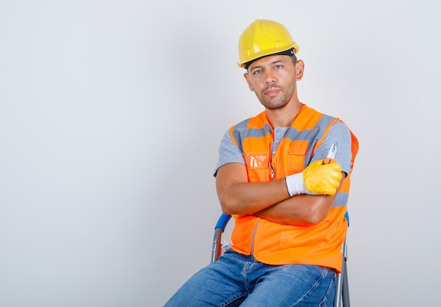 Costruttore maschio in uniforme, jeans, casco, guanti seduti con le braccia incrociate, vista frontale.