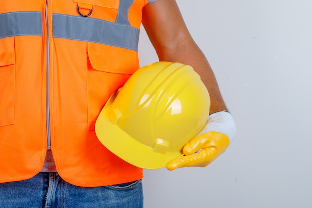 Costruttore maschio in uniforme, jeans, guanti che tengono il casco in mano, vista frontale.