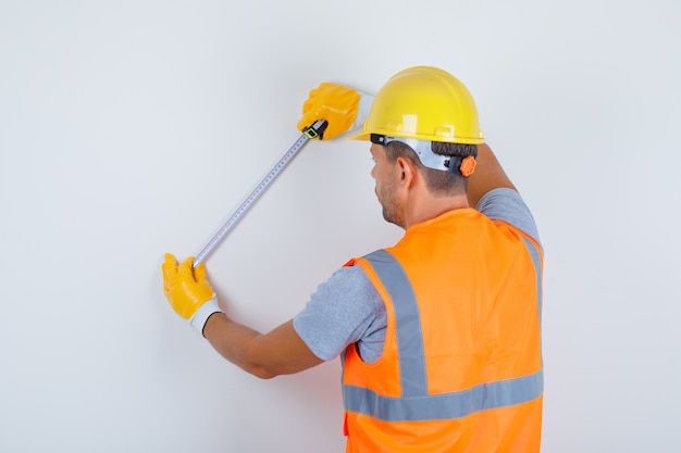 Costruttore maschio utilizzando nastro di misurazione sulla parete in uniforme, casco, guanti e guardando occupato, vista posteriore.