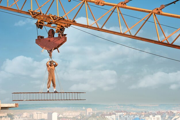 Costruttore muscoloso con torso nudo in piedi su una costruzione in ferro in alto e tenuto da funi. Uomo che indossa un cappello e abiti da lavoro che guarda l'obbiettivo. Cielo azzurro con nuvole sullo sfondo.