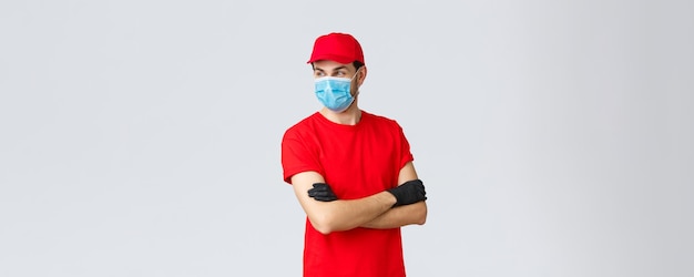 Covid-19, auto-quarantena, shopping online e concetto di spedizione. Corriere fiducioso in uniforme rossa, guanti protettivi e maschera facciale, braccia incrociate in piedi, distogliere lo sguardo, pronto a prendere i tuoi ordini