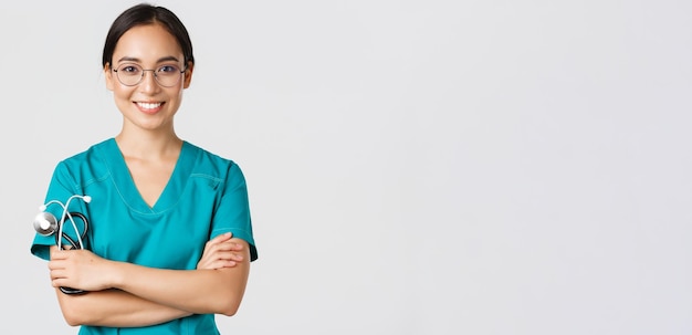 Covid-19, malattia da coronavirus, concetto di operatori sanitari. Primo piano di una dottoressa professionista sicura di sé, infermiera con gli occhiali e scrub in piedi su sfondo bianco, braccia incrociate.