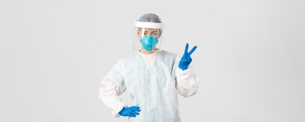 Covid-19, malattia da coronavirus, concetto di operatori sanitari. Sassy dottoressa asiatica, medico o laboratorio tecnologico in dispositivi di protezione individuale che mostra il segno di pace, in piedi su sfondo bianco.