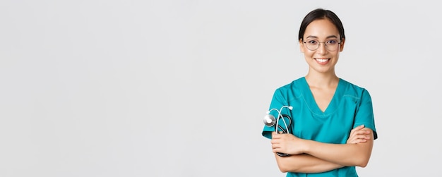 Covid-19, malattia di coronavirus, concetto di operatori sanitari. Primo piano di una dottoressa professionale sicura, un'infermiera con gli occhiali e uno scrub in piedi su sfondo bianco, braccia incrociate.