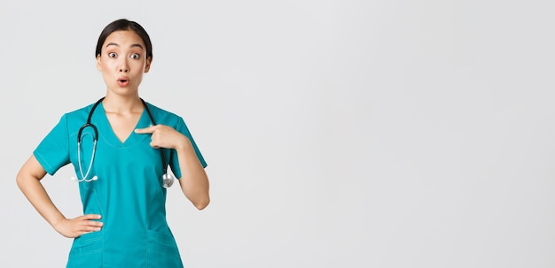Covid-19, operatori sanitari, concetto di pandemia. Medico femminile asiatico sorpreso e teso un'imboscata, infermiera in camice che indica se stessa, battendo il nome o la scelta, in piedi su sfondo bianco.