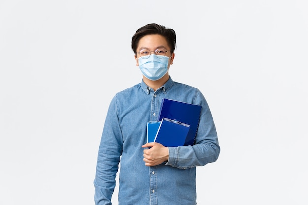 Covid-19, prevenzione del virus e distanza sociale al concetto universitario. Il giovane insegnante asiatico bello, l'insegnante maschio o lo studente in maschera medica portano i taccuini per la lezione, fondo bianco.