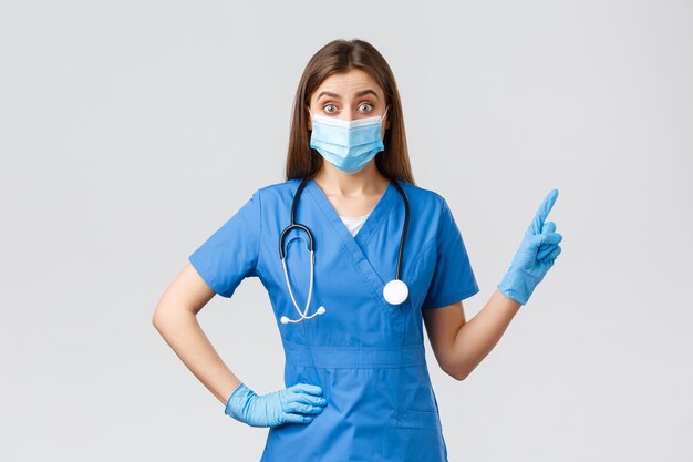 Covid-19, prevenzione di virus, salute, concetto di operatori sanitari. La dottoressa incuriosita ed eccitata, l'infermiera in camice blu e la maschera medica informano i pazienti, puntando le dita nell'angolo in alto a destra