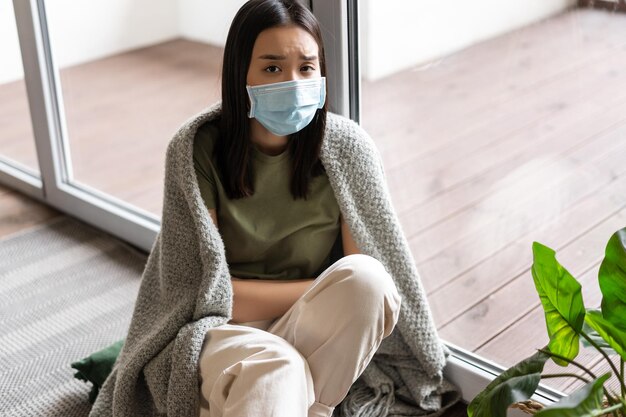 Covid e concetto di pandemia triste ragazza asiatica malata in maschera medica autoquarantena a casa guardando o...