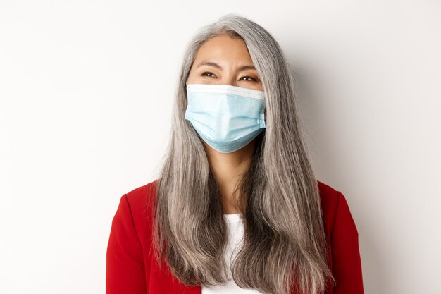 Covid, pandemia e concetto di business. Primo piano di felice imprenditrice asiatica con i capelli grigi, indossando maschera medica e sorridente, guardando a sinistra con la faccia allegra, sfondo bianco.