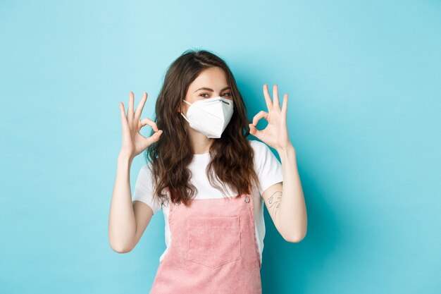 Covid, salute e concetto di pandemia. Molto bene. Giovane donna di supporto che indossa un respiratore medico e mostra segni di approvazione in segno di approvazione, loda indossando maschere per il viso in pubblico, sfondo blu.