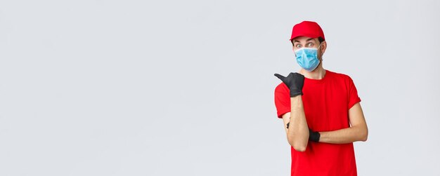 Covid19 selfquarantine shopping online e concetto di spedizione Eccitato uomo di consegna sorpreso con maschera facciale uniforme rossa e guanti che punta a sinistra verso la promozione che mostra i bonus o l'applicazione ai clienti