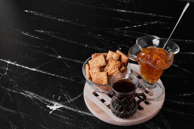 Cracker croccanti in una tazza di vetro sulla superficie nera con un bicchiere di tè e confettura.