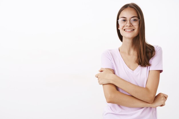 Creativa e intelligente bella donna in occhiali roung e camicetta tenendo la mano sul braccio in una timida postura insicura sorridente sensazione di non sicurezza durante il primo giorno di lavoro sul muro bianco