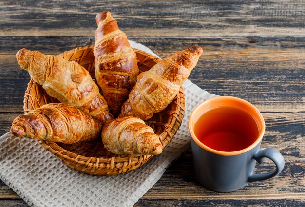 Croissant con la tazza di tè in un canestro sull'asciugamano di cucina e di legno, vista dell'angolo alto.