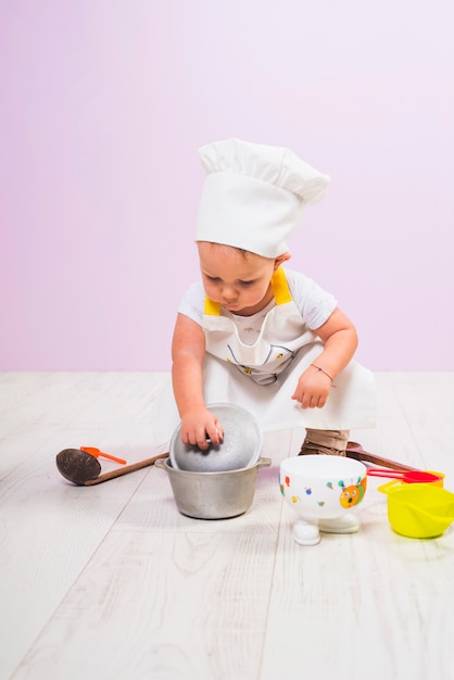 Cucini il bambino che si siede con gli utensili della cucina sul pavimento